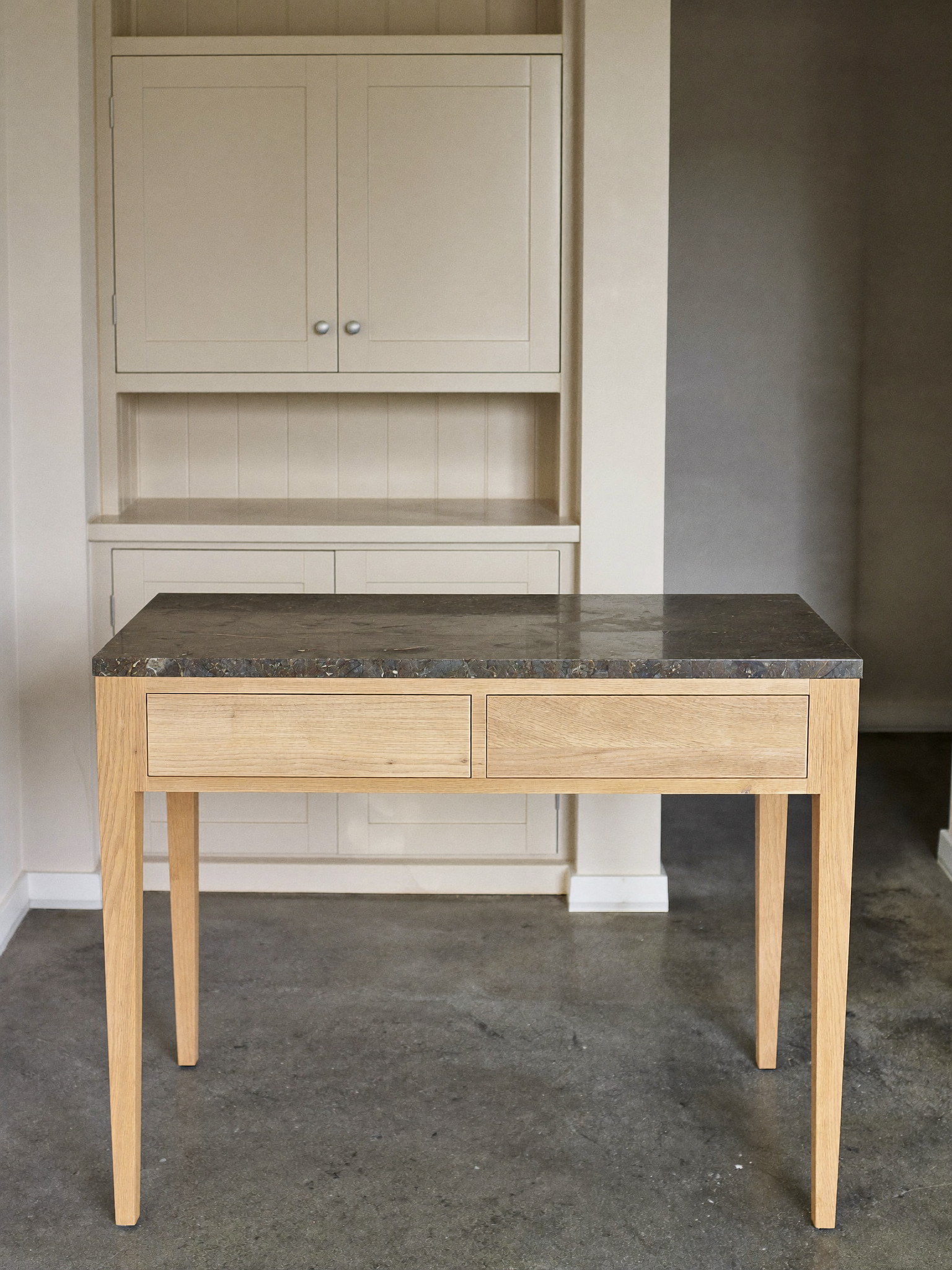 A slim freestanding white oak work table with a charcoal soapstone top in a compact renovated city kitchen, simple square legs, plain drawer faces with no