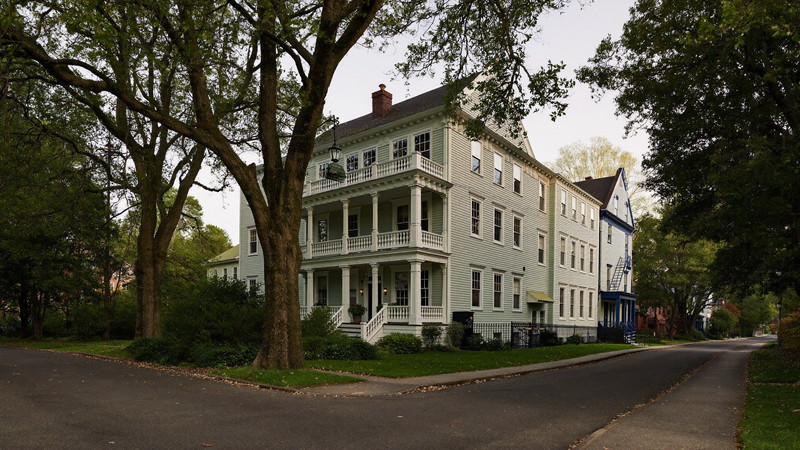 A leafy Cambridge Massachusetts residential street in soft evening light: painted wooden triple-decker homes with layered rear porches and detailed trim