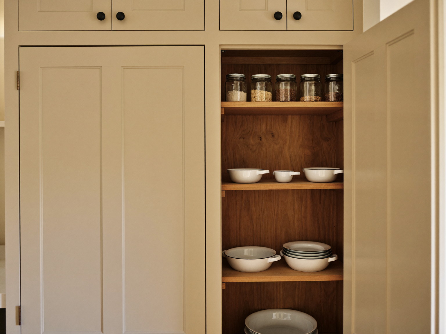 Floor-to-ceiling pantry joinery in a renovated Cambridge kitchen: putty-painted inset doors open to white oak shelves holding plain white ironstone and clear