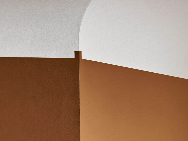 Macro photograph of a razor-sharp masked paint line where a warm clay-toned wall meets a bright white plaster ceiling, the edge perfectly straight and clean