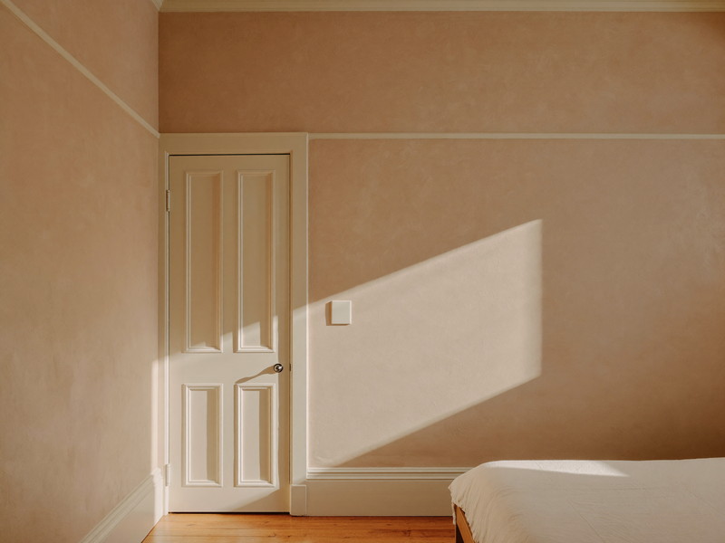A repainted bedroom in an older Cambridge condominium: soft warm clay-putty walls, bright white trim and paneled door, restored plaster smooth in raking light