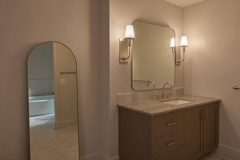 A calm finished suburban bathroom in soft evening light: satin nickel sconces glowing beside an arched mirror, warm taupe vanity with a completely bare granite top