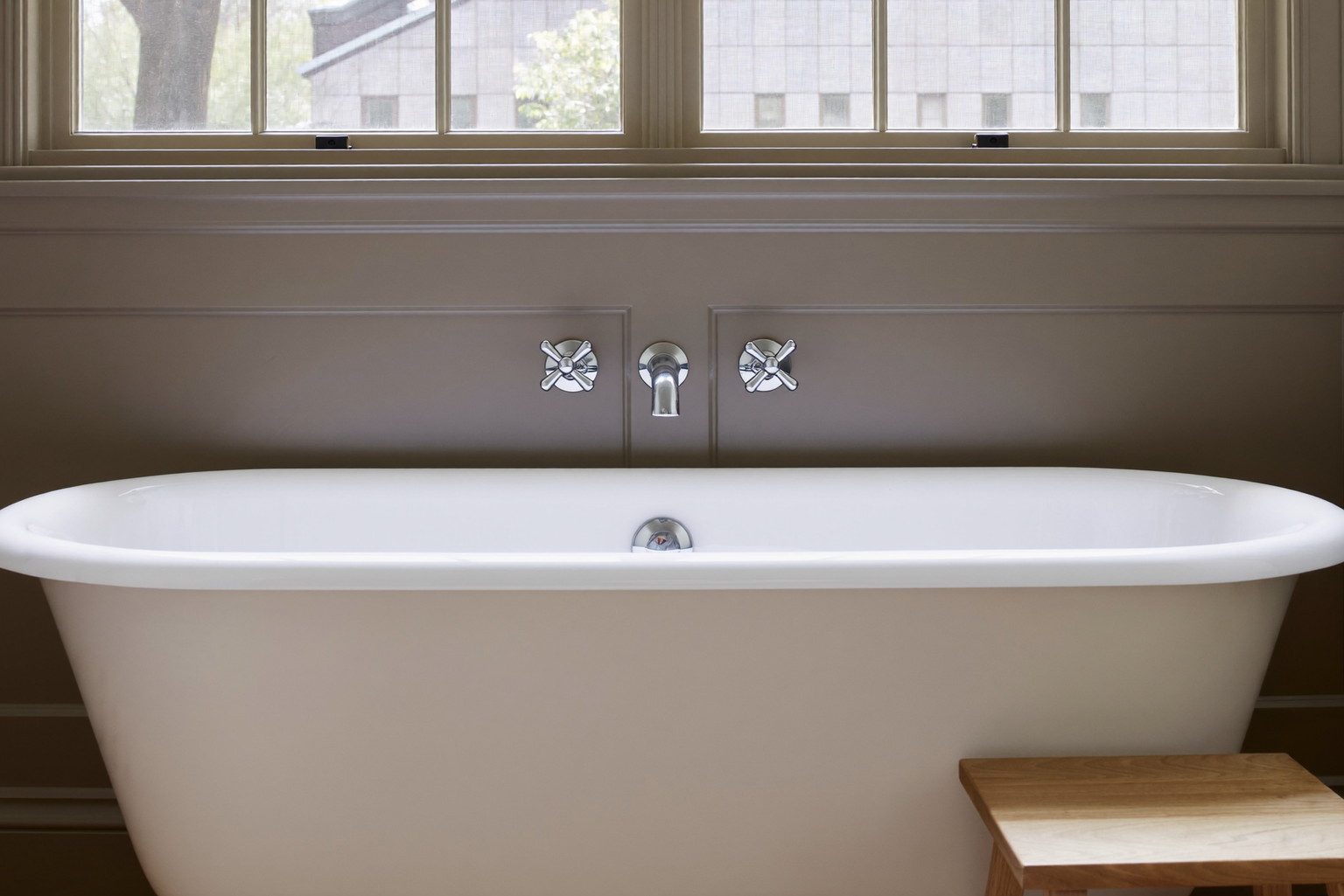 A renovated bathroom in an established Framingham colonial: white freestanding tub beneath a window with muntins, warm taupe wainscot