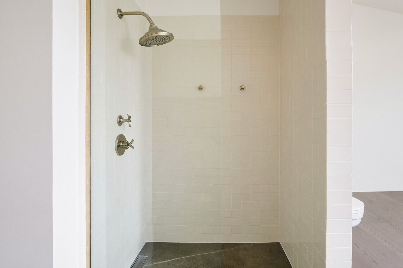 A walk-in shower behind a clear glass panel in a renovated suburban bathroom: soft cream rectangular tile, satin nickel rain head and controls, honed gray floor