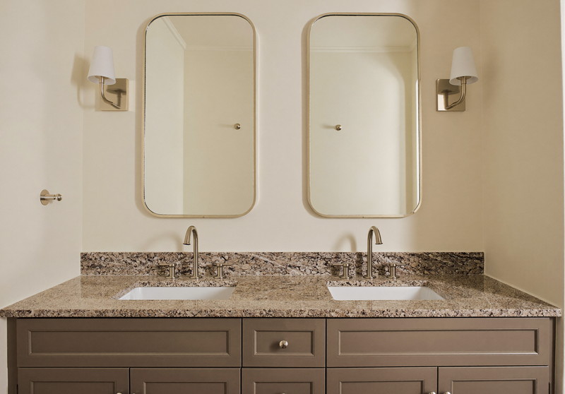 A double warm taupe vanity with honed granite top under two softly rounded nickel-framed mirrors, satin nickel sconces, soft cream walls, comfortable suburban bathroom