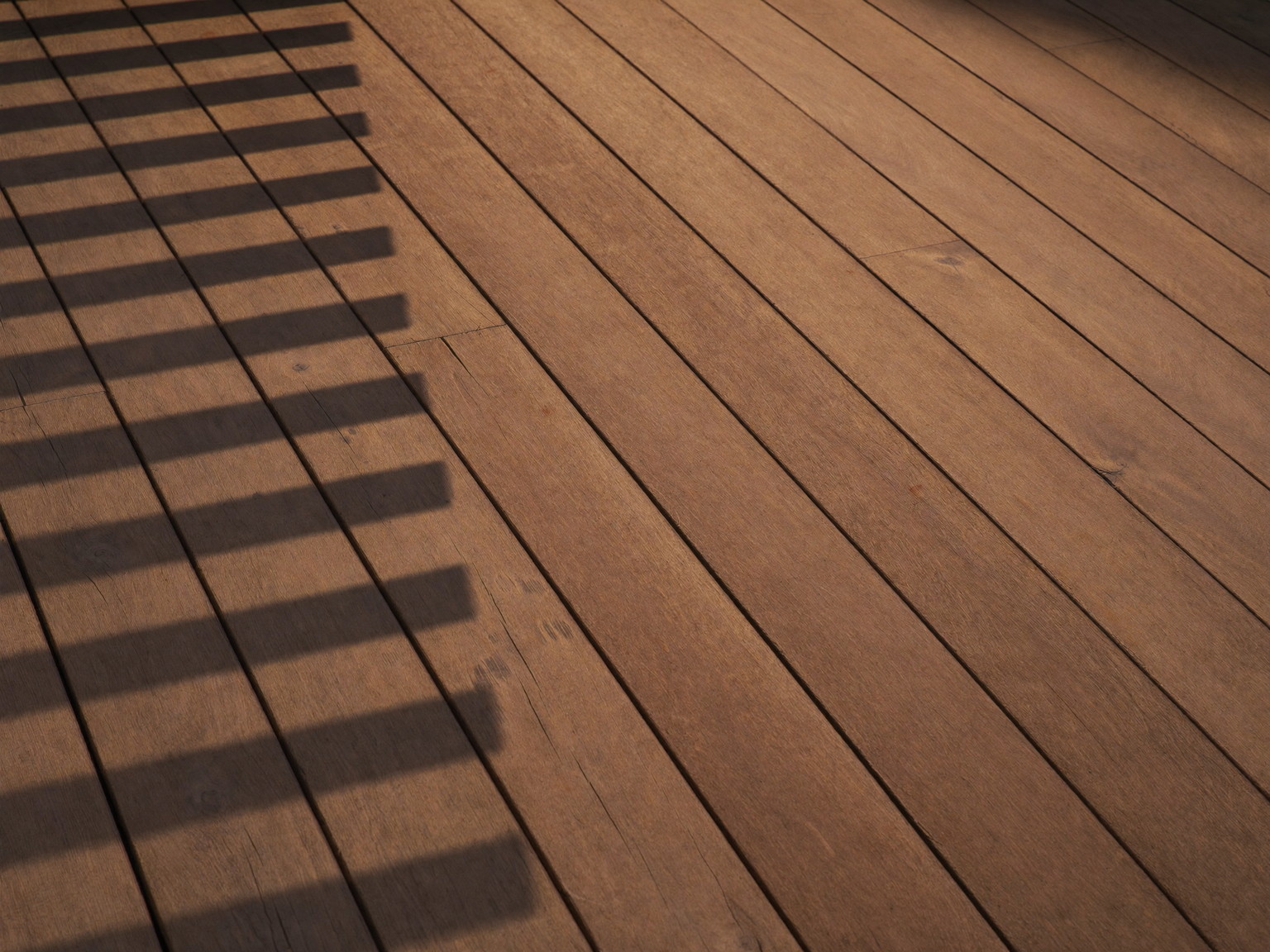 A freshly stained warm-brown deck surface drying in afternoon light: smooth even boards with a soft sheen, crisp black baluster shadows falling across the grain
