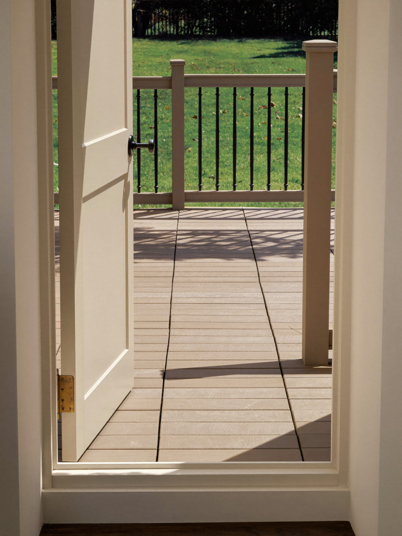 Tall photograph through an open cream back door onto a rebuilt deck: pale warm decking boards, taupe posts with black balusters, deep green lawn beyond, bright day