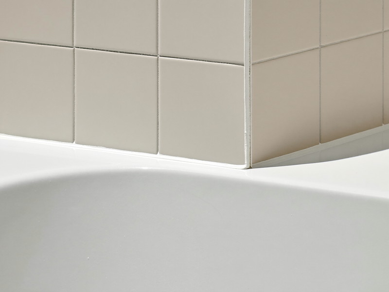 A brand-new bright white bathtub edge meeting freshly grouted soft cream wall tile, crisp bright grout lines, the joint finished with a smooth clean bead