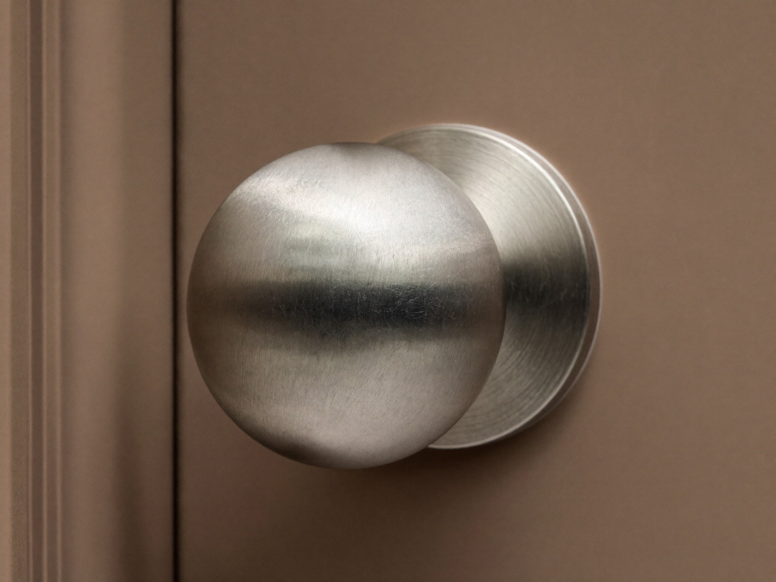 Macro photograph of a classic satin nickel round door knob, a smooth plain ball-shaped knob on a simple round rosette, mounted on a warm taupe paneled door