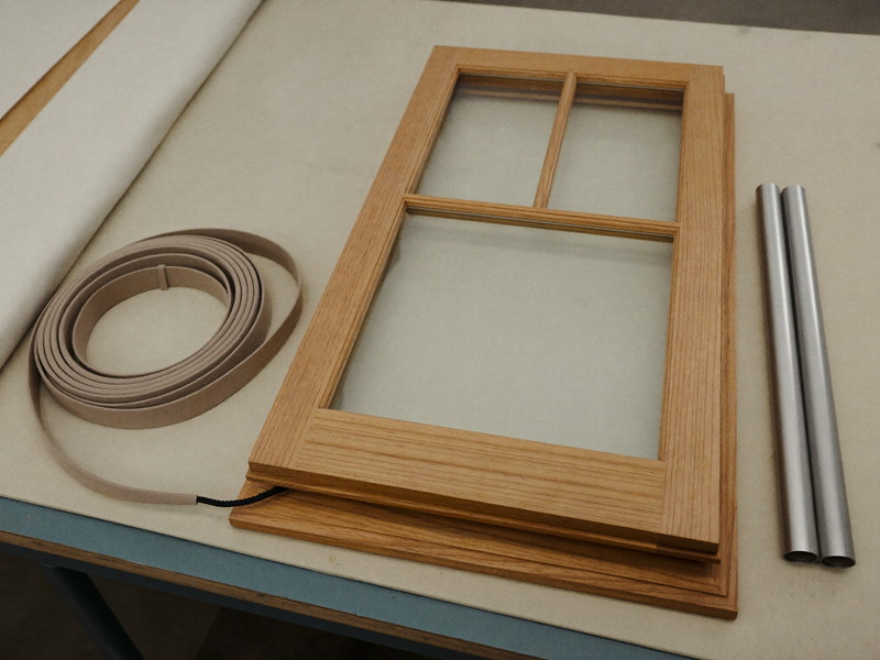 A wooden double-hung window sash lying flat on a padded workbench, a neat coil of soft cotton sash tape and two plain smooth steel cylinders resting beside the sash