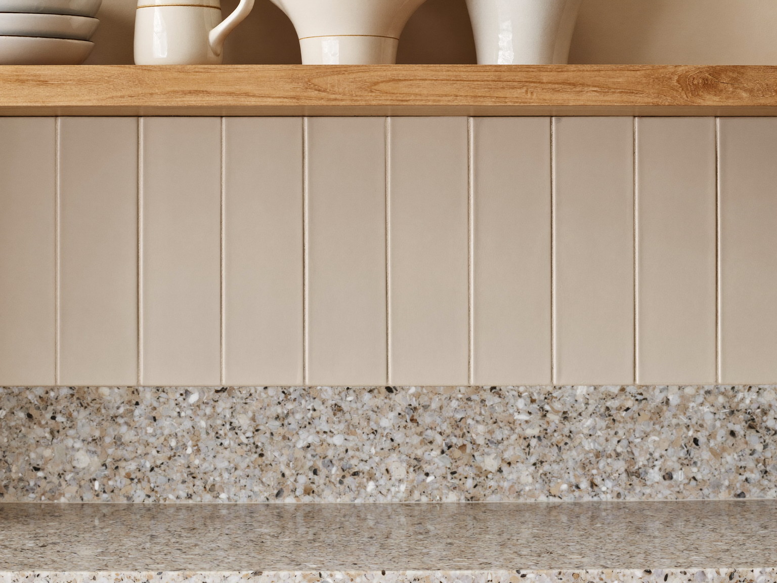 Macro photograph of a honed speckled granite counter band meeting soft cream beadboard paneling, a slim hickory shelf above holding plain white ceramics