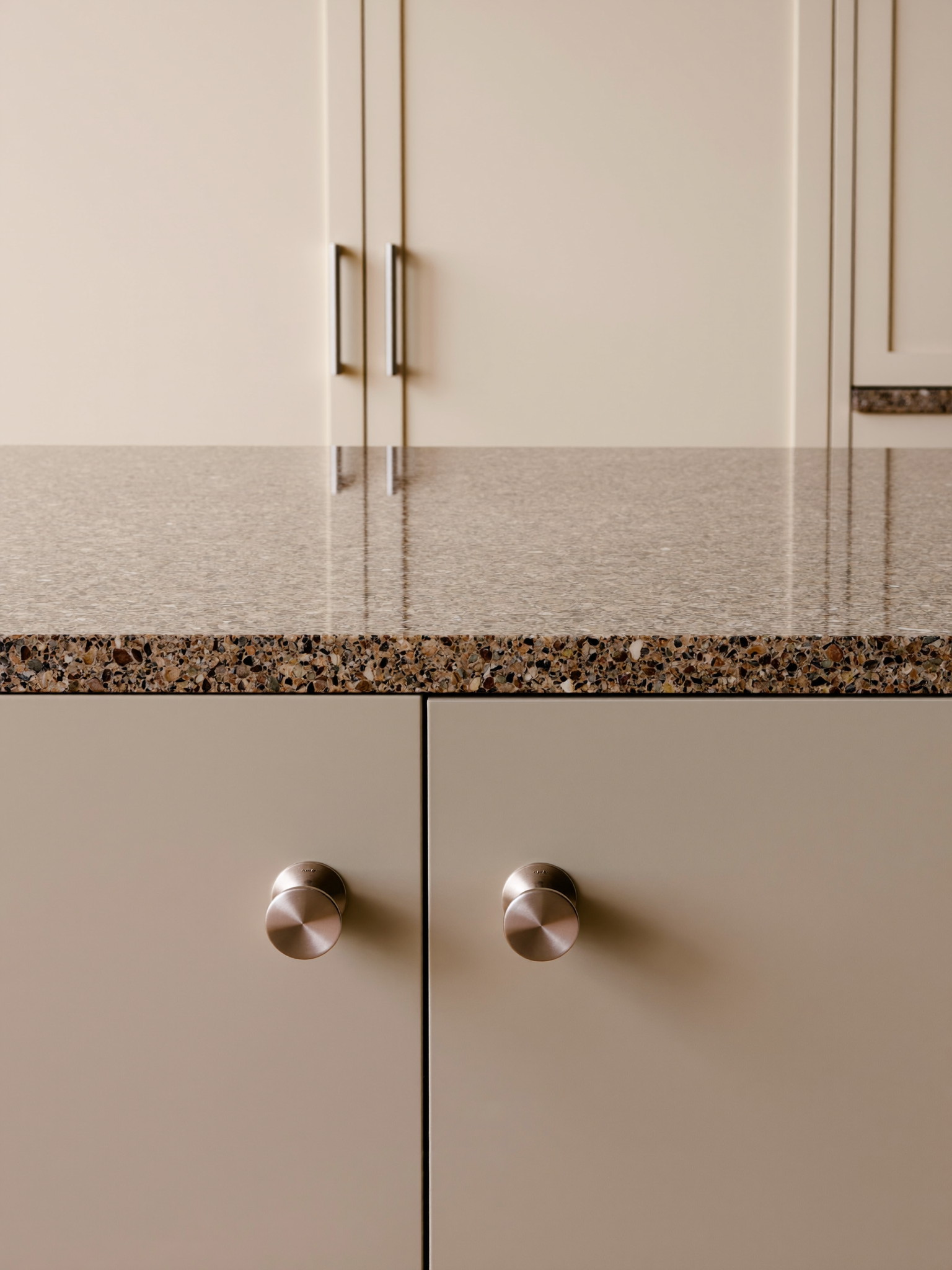 A warm taupe kitchen island with a thick honed speckled granite top, satin nickel bar pulls with smooth faces, soft cream shaker cabinets behind, natural light