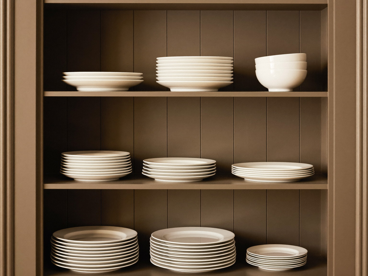 Inside an established colonial's pantry: warm taupe painted shelves holding only stacked plain white dinner plates and plain white bowls