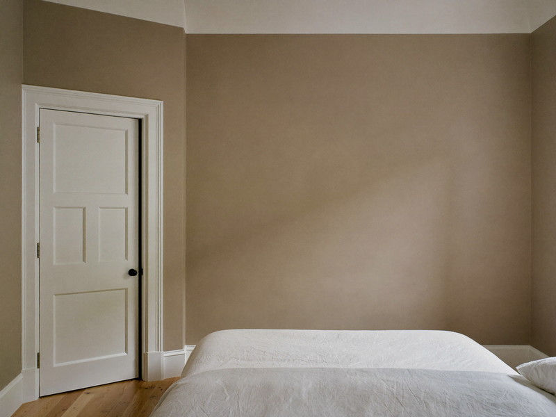 A repainted bedroom in an established Framingham colonial: soft mushroom-taupe walls, bright white trim and paneled door, smooth restored plaster in raking light