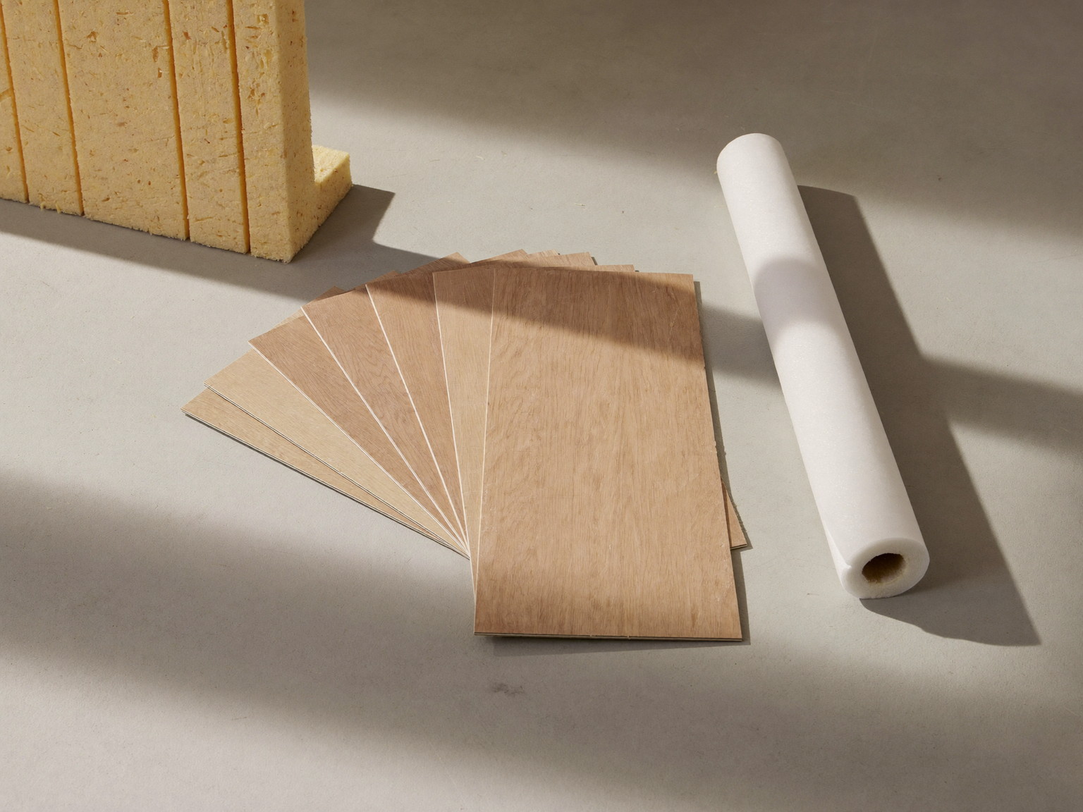 Material still life: oak-tone vinyl plank samples, rigid insulation corner, moisture barrier roll, and a blank-faced moisture meter on concrete