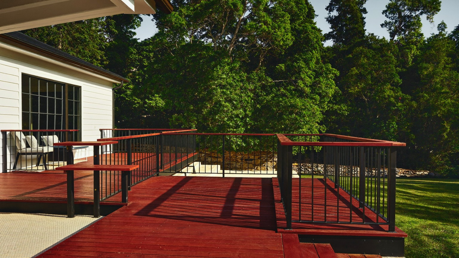 Mahogany-toned deck with black metal railings wrapping a white New England colonial, summer maples and a stone wall beyond
