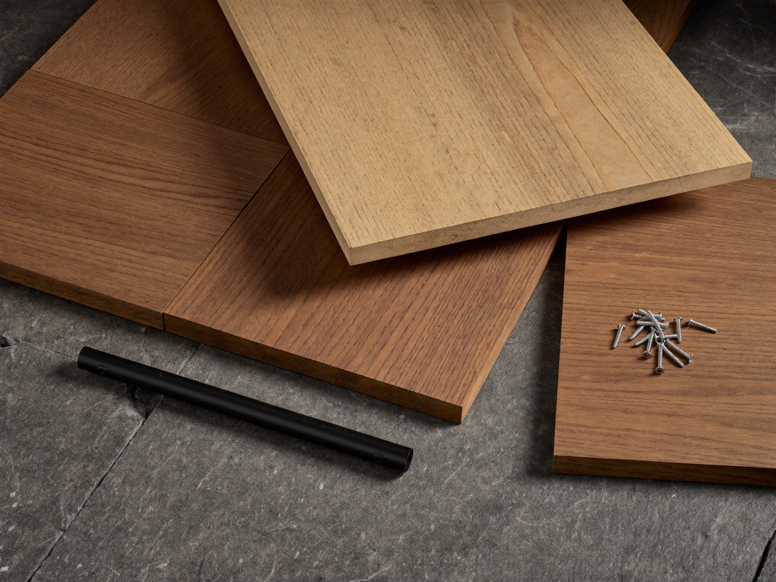 Material still life: cedar and charcoal composite deck boards, black railing tube, stainless fasteners, and a sealed finish sample on slate