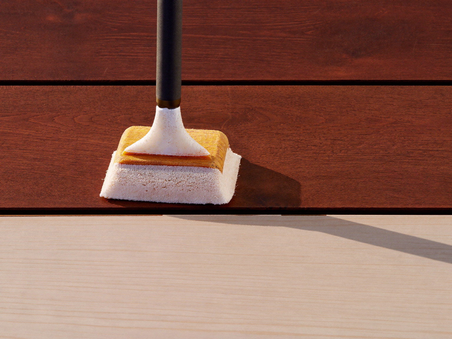 Deck staining in progress: an applicator pad mid-stroke where rich sealed boards meet dry unfinished decking