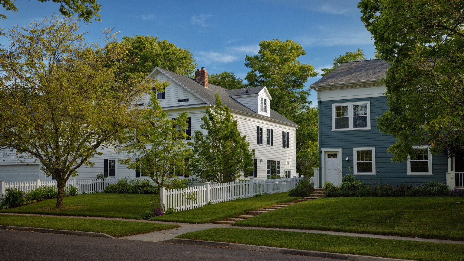 Quiet Massachusetts residential street in summer with a white Colonial and slate-blue Cape among oaks and maples