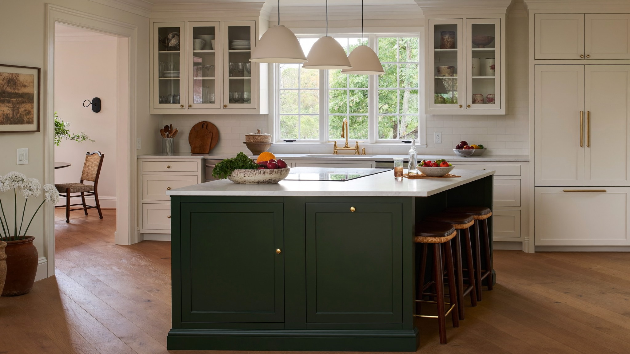 Deep view through a remodeled New England kitchen: forest-green island with brass pulls, white shaker perimeter, and warm oak floor