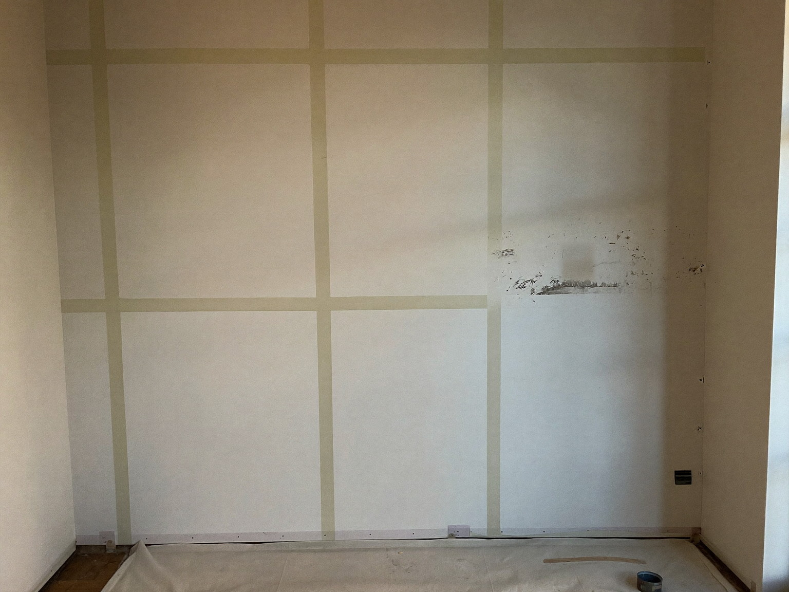Older plaster wall mid-preparation: sanded joint compound patches and spot-primed repairs above a floor protected with a canvas drop cloth