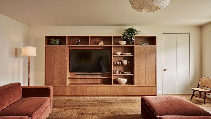 A finished suburban lower-level family room: warm ivory walls, walnut built-in media wall with closed plain cabinet doors and open shelves holding plain