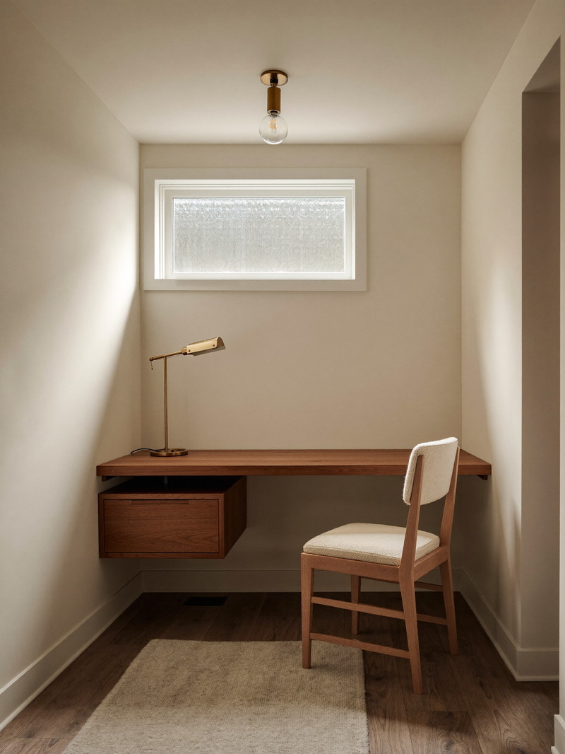 A compact finished basement office nook: warm ivory walls, low ceiling, one small high awning window near the ceiling glowing with soft frosted daylight and