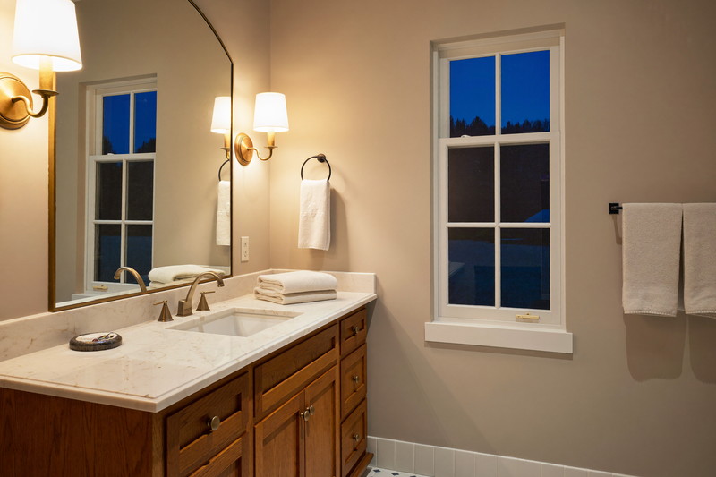 A calm finished suburban bathroom at dusk: brass sconces lit beside an arched mirror, cherry vanity, white quartzite top, white zellige tile glowing warm