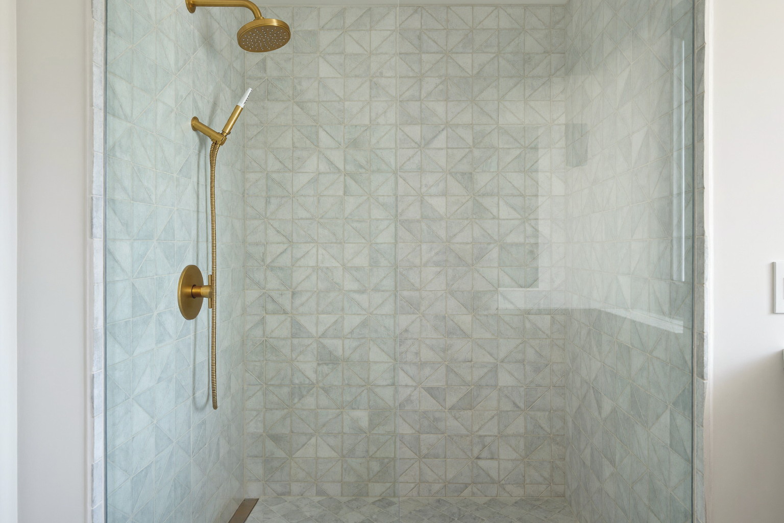 Wide photograph of a walk-in shower behind clear frameless glass in a renovated suburban bathroom: white zellige-style tile walls, pale gray limestone-look