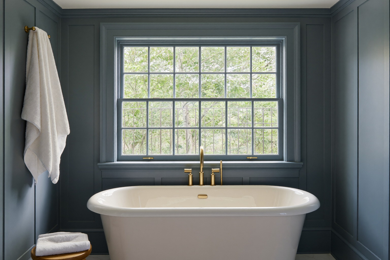 A renovated bathroom in an established New England home: white freestanding tub under a wide window with muntins, slate-blue paneled walls, warm white