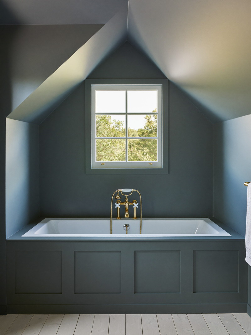 A renovated bathroom in an established New England cape: slate-blue walls under a sloped dormer ceiling, white tub with brushed brass fixtures, white oak