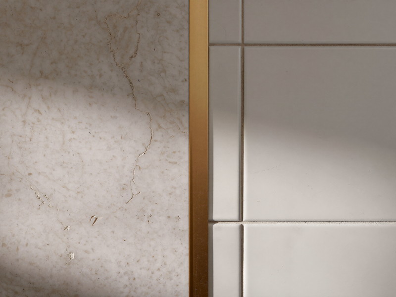 Macro photograph where honed white quartzite meets white zellige-style ceramic tile with a crisp caulk line, slim brushed brass trim strip between, beautiful