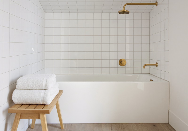 A white tub-and-shower alcove newly rebuilt with white square tile to the ceiling: one single wall-mounted brushed brass tub spout on the back wall with one