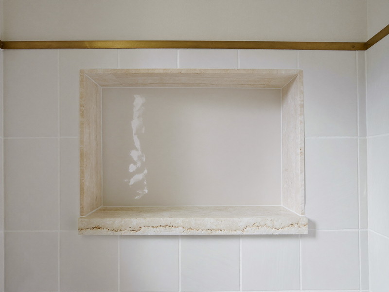 Detail of a walk-in shower in an established Hopkinton-style remodel: white handmade-look tile, recessed niche with a single honed stone shelf, brushed brass