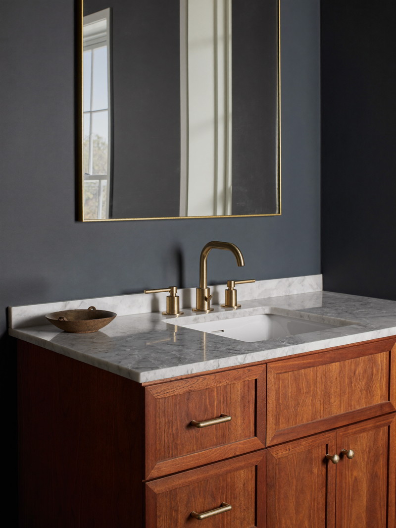 A furniture-style cherry wood bathroom vanity with a honed white quartzite top and brushed brass faucet with smooth lever handles, arched mirror in a thin