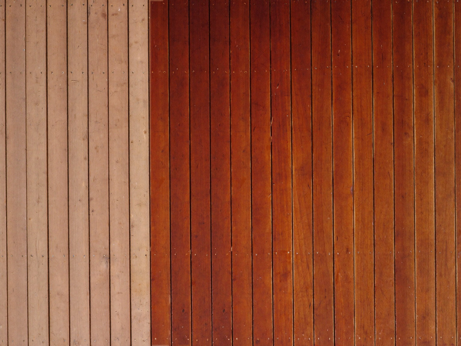 Top-down photograph of a cedar deck mid-restaining: rich warm semi-transparent stain covering the right half of the boards, meeting bare sanded wood at one