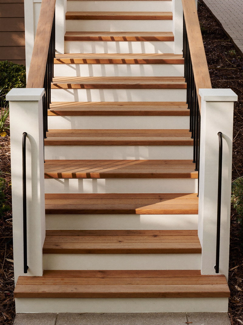 Cedar deck stairs descending from an established suburban home: warm cedar treads, white painted risers and posts, black metal balusters, each stair landing