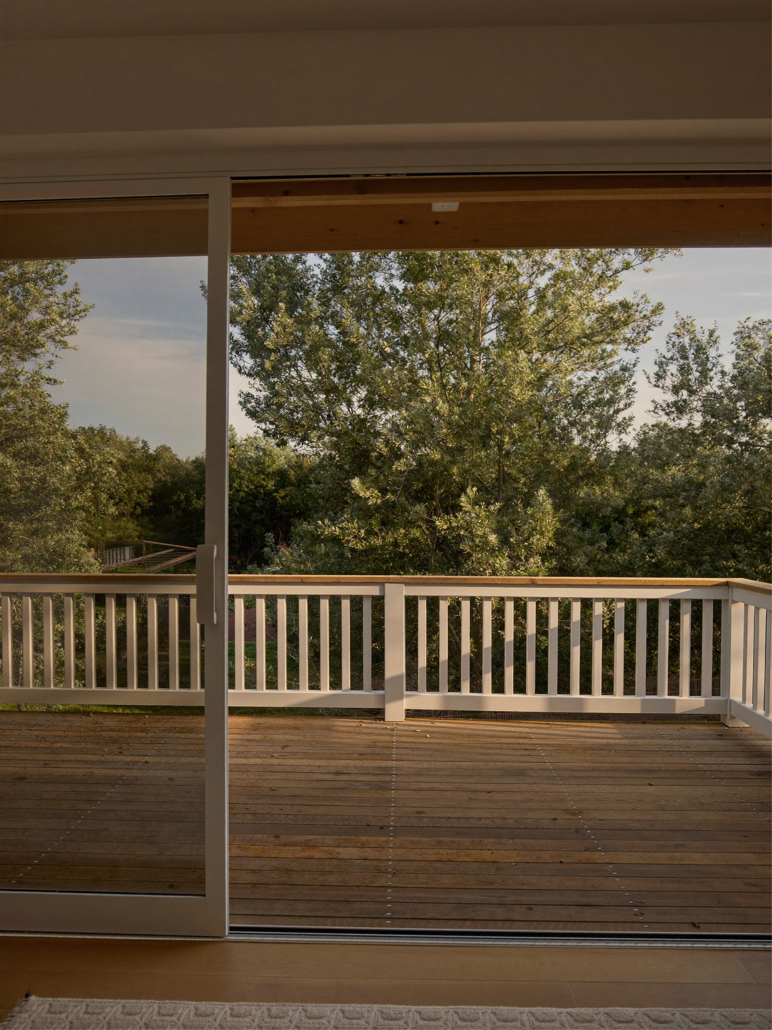 View through a wide sliding glass door from a warm family room onto a cedar deck with white and black railing and a wooded suburban back yard beyond, late