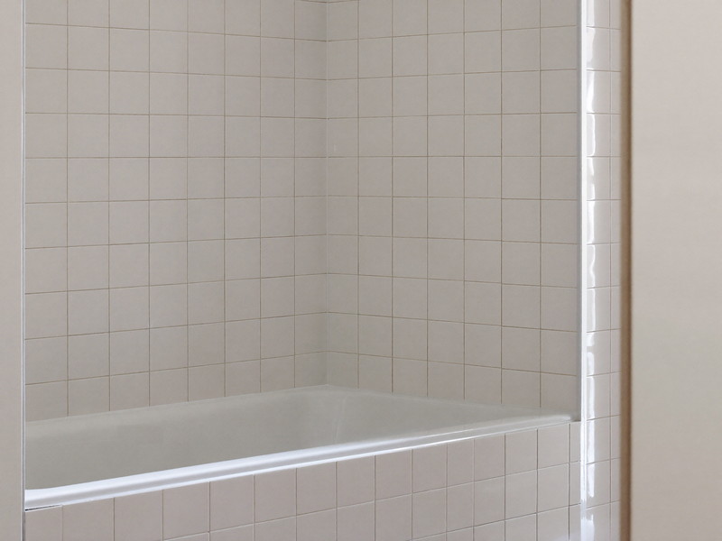 Bright corner of a freshly renovated bathroom: glossy white bathtub meeting square white ceramic tile walls, crisp clean tile joints, everything dry and