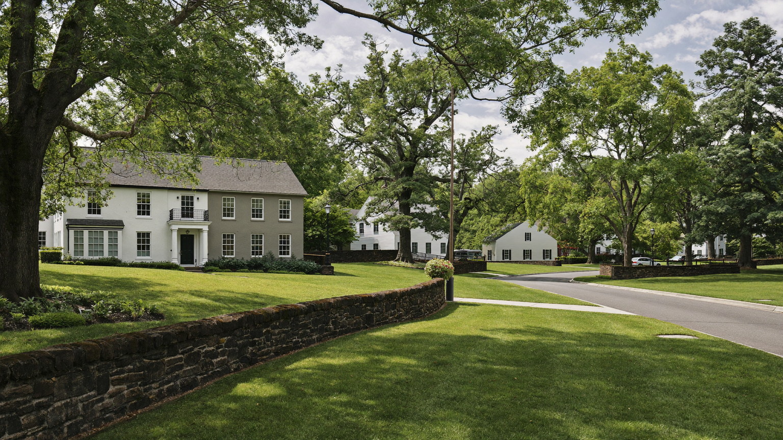 A quiet winding tree-lined Hopkinton Massachusetts style suburban road in summer: white and gray colonial and cape homes set back on green lawns, low