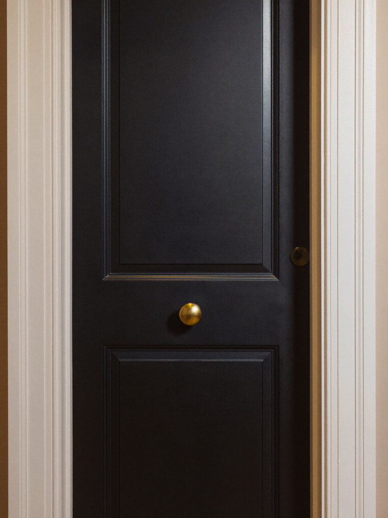 An interior paneled door freshly painted in soft black satin enamel with crisp edges against white casing, plain unmarked brass knob, warm hallway light