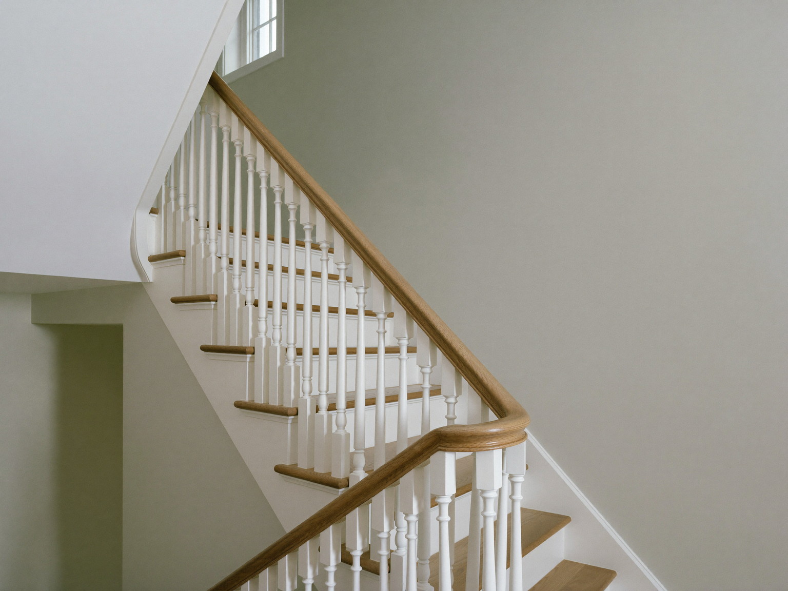 A repainted colonial stair hall: soft greige walls, bright white trim, painted white balusters with an oak handrail, oak treads, tall window at the landing