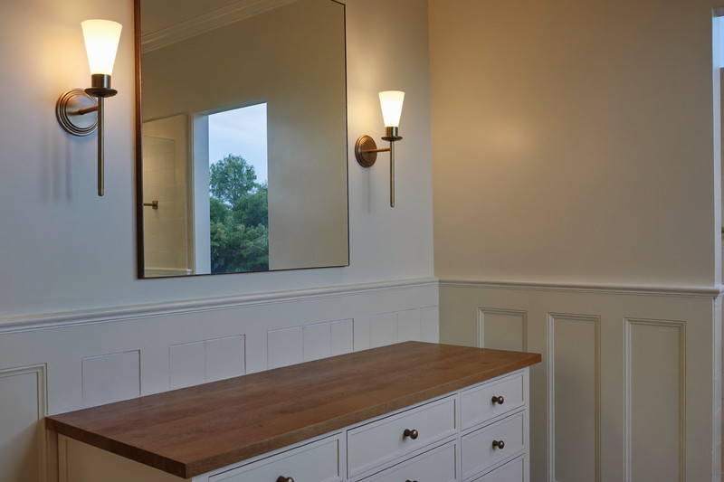A calm finished colonial bathroom in soft evening light: brushed pewter sconces glowing beside a rectangular mirror, ivory vanity with a completely bare maple top