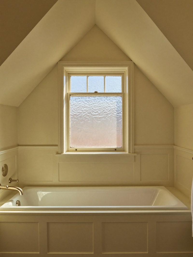 A renovated bathroom under the sloped ceiling of a Lexington antique cape: white tub with a completely bare rim, ivory paneling, brushed pewter fixtures
