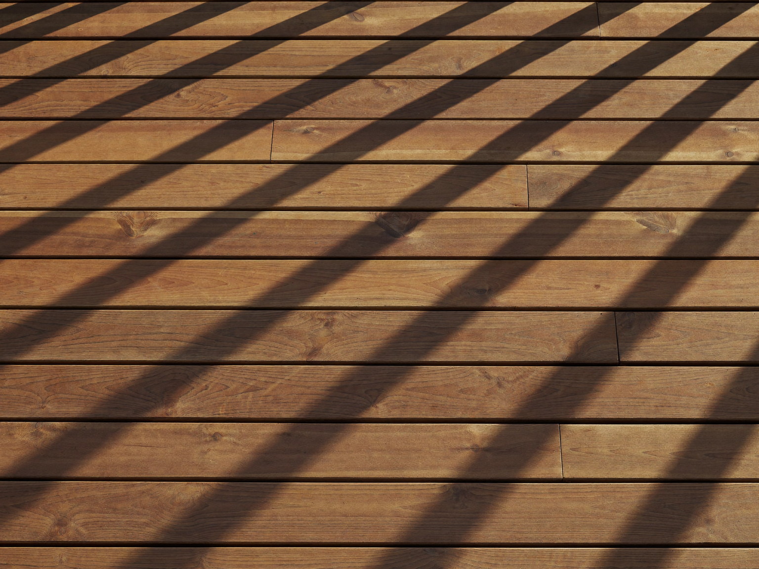A freshly oiled cedar deck surface drying in warm afternoon light: smooth even boards with a soft sheen, crisp black baluster shadows falling across the grain