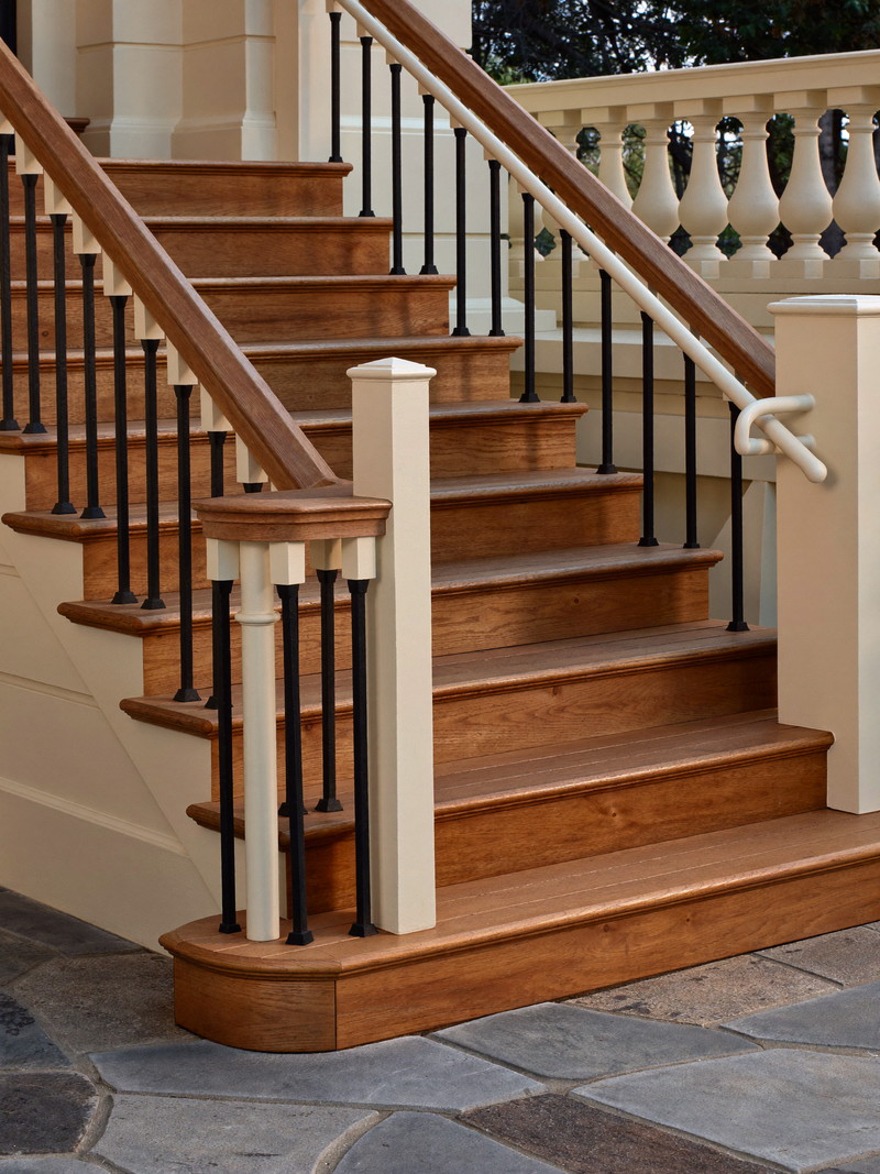 Rebuilt deck steps on a historic colonial: warm cedar treads and risers, ivory posts with black metal balusters, simple handrails, stone paving below, afternoon light