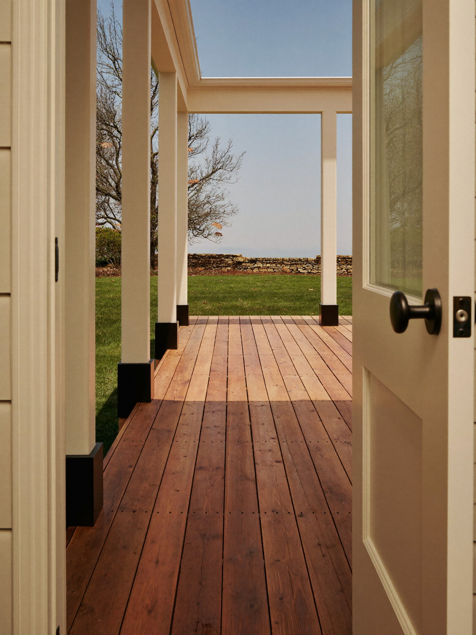 Tall photograph through an open ivory back door onto a rebuilt cedar porch deck: warm decking boards, ivory posts, deep green lawn and a low fieldstone wall beyond