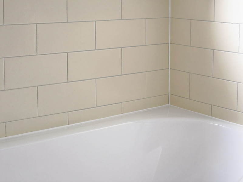 A clean bright white bathtub edge meeting ivory subway wall tile, the joint freshly finished, smooth and tidy, completely bare tub edge, soft daylight