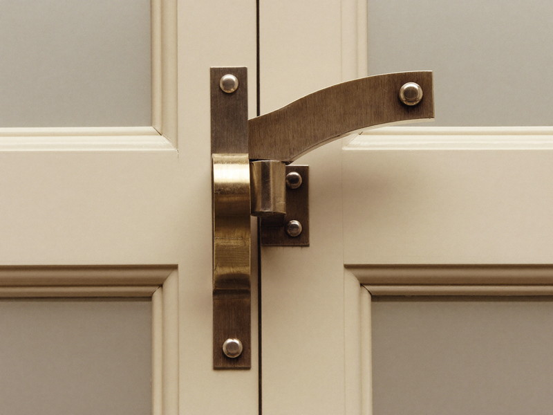 Close view of a traditional aged-bronze window sash lock with a curved swing arm where two ivory painted wooden sashes meet, smooth plain metal, crisp paint, soft light