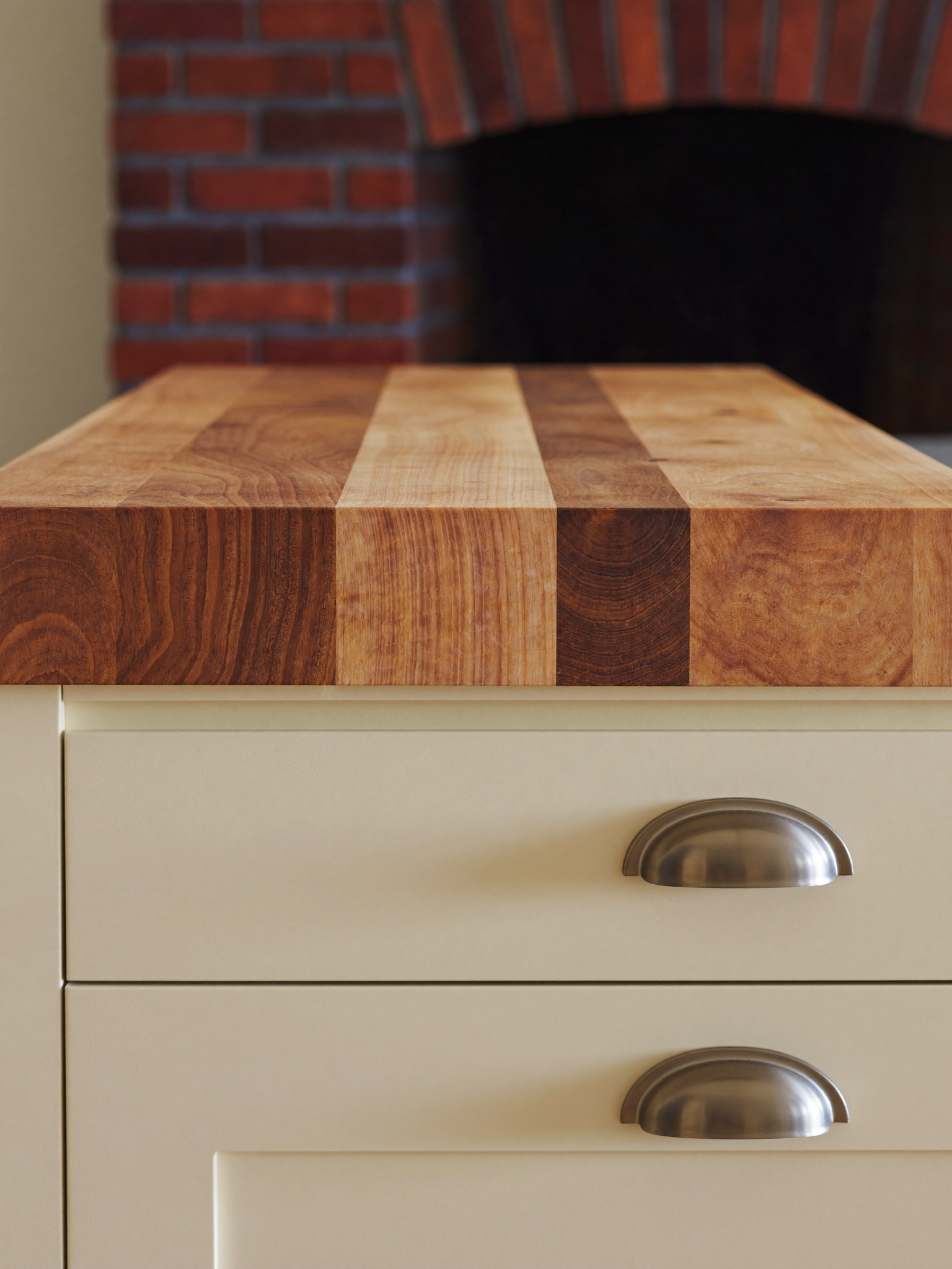 An ivory kitchen island with a thick warm maple butcher-block top with visible wood grain, brushed pewter cup pulls with smooth faces